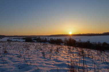 Altın bir günbatımında karla kaplı tepelerin ve tarlaların panoramik manzarasını içeren manzaralı kış manzarası. Parlak güneş ışınları pırıl pırıl karı ve kuru otları aydınlatarak soğuk bir akşamda huzur, sessizlik ve doğal bir uyum ortamı yaratır.