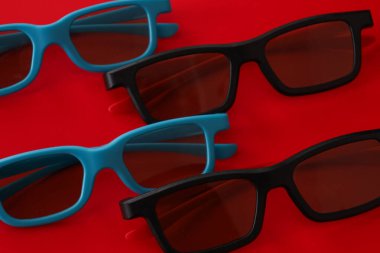 Siyah ve cyan çerçeveli dört adet şık üç boyutlu gözlük parlak kırmızı bir arkaplanda rastgele düzenlenmiştir. Karşıt renk paleti ve minimalist kompozisyon heyecan verici dünyayı sembolize eden modern bir görsel görüntü oluşturur.