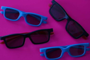 Mavi ve siyah çerçeveli dört çift modern 3D sinema gözlüğü parlak doymuş pembe arka planda yatar. Film aksesuarları ile bu minimalist kompozisyon zıt renkler ve geometrik düzenleme yoluyla dinamik ve şık bir görünüm yaratır..