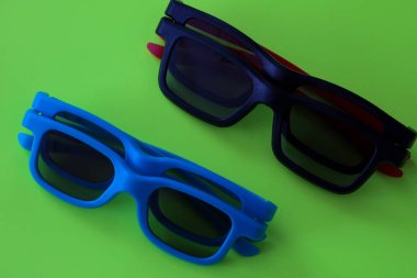 İki adet katlanmış mavi ve siyah renkli 3D gözlük parlak yeşil bir arka planda paralel olarak düzenlenmiştir. Minimalist tasarım ve doymuş neon arkaplan çarpıcı bir görsel karşıtlık yaratır,