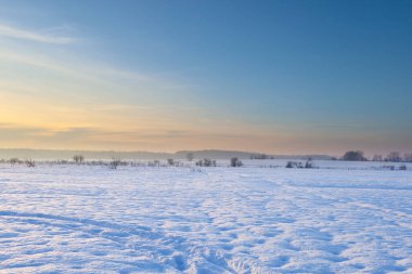 Karla kaplı geniş bir tarla, açık mavi gökyüzüne karşı yükselen kış güneşinin yumuşak ışınlarının altında parıldıyor. Ufukta yumuşak altın ışıkla bu sabah manzara sınırsız bir huzur hissi yaratıyor.