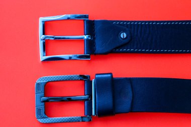 Bir çift şık erkek siyah deri kemeri farklı metal toka dizaynları ile canlı kırmızı arka planda.