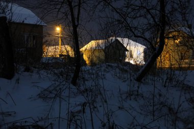 Sıcak geceyi aydınlatan sıcacık kır evleri, karlı alacakaranlıkta kalın bir ağaç dallarının arasından dikiz dikiyor..
