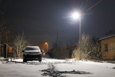Yerleşim alanındaki sessiz, karlı bir cadde parlak bir sokak lambasıyla aydınlatılır. Uzun gölgeler ve taze kar üzerinde büyülü bir parıltı yaratır..