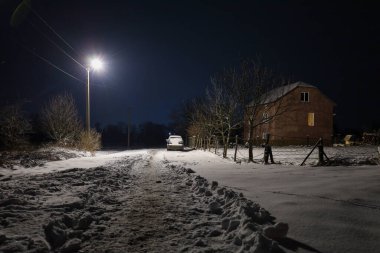 Karlı bir yolu aydınlatan parlak bir sokak lambası derin bir gece gökyüzünün altındaki ıssız bir eve götürüyor. Soğuk mavi akşam ile penceredeki sıcak ışık arasındaki zıtlık, karlı banliyölerde huzur ve huzur ortamı yaratıyor..