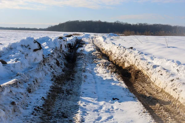 Güneşli bir kış gününde, karlı bir arazide ağır makinelerin yol aldığı derin ve çamurlu bir yol. Karla karışmış toprak ve temiz ayak izleri temiz bir gökyüzünün altında uzak bir orman kuşağına uzanan zıt bir yol yaratır..