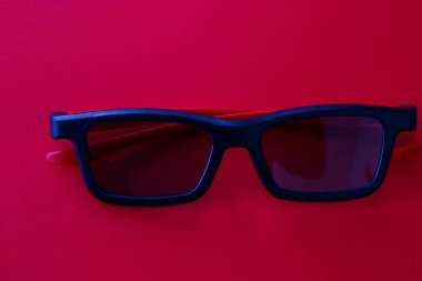 Kırmızı şakaklı siyah 3D gözlükler zengin kırmızı arka planda yatar ve şık bir tek renkli etki yaratır. Derin tonlar ve minimalist kompozisyon, sinema ve dijital eğlencenin modern estetiğini vurgular..