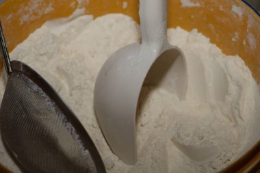 Büyük bir portakal kabında beyaz buğday ununun Macro fotoğrafı. Una plastik bir kepçe batırılır ve yakınlarda bir metal elek bulunur. Bu da pişirme ya da pişirme için hazırlanma sürecini gösterir..