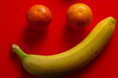 Olgun sarı bir muz ve iki turuncu mandalinanın yaratıcı kompozisyonu parlak kırmızı bir arka planda neşeli bir gülümseme yaratır..