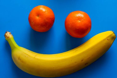 Parlak sarı bir muz ve iki turuncu mandalinadan oluşan bir kompozisyon, zengin mavi arka planda gülümseyen bir yüz oluşturur..