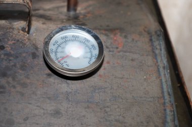 Çift ölçekli yuvarlak analog termometre, endüstriyel ekipmanların kaba metal yüzeyine monte edilir. Açık gösterge sıcaklığı Celsius derecesinde gösterir. Pas ve kaynak izlerini gösteren koyu bir arkaplan ile çelişir..