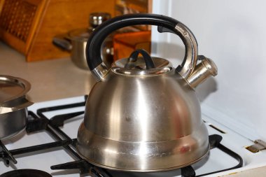 Islık çalan ve siyah saplı klasik bir çelik çaydanlık sıcak bir mutfak içinde beyaz bir gaz sobasının üzerinde oturur. Parlak metal yüzey ışığı yansıtarak, ev hayatının, sabah çayının ya da kahve hazırlığının pratik bir resmini oluşturur.,