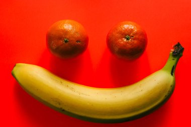 İki mandalina ve canlı kırmızı arka planda olgun sarı bir muz kullanılarak yaratılmış gülen bir yüzün yaratıcı ve olumlu bir görüntüsü. Bu neşeli meyve kompozisyonu iyi havayı simgeliyor.,