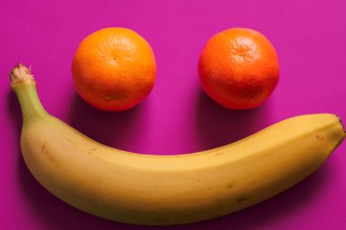 Gülümseyen yüz şeklindeki olumlu bir kompozisyon, canlı mor arkaplanda iki turuncu mandalina ve olgun sarı bir muzdan meydana gelir..