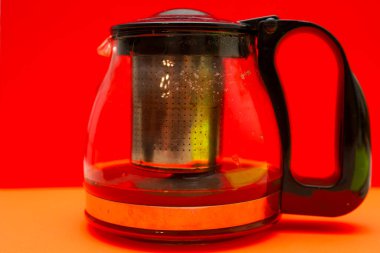A glass teapot with a metal infuser and hot beverage stands out against a rich red and orange background. The minimalist design emphasizes the transparency of materials,