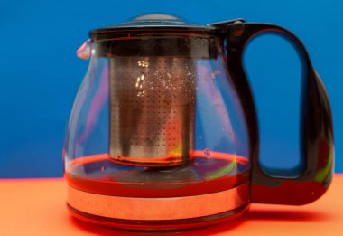 A glass teapot with a metal infuser is filled with rich dark tea, creating a vivid contrast against a blue and orange background.