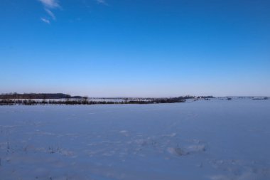 Uçsuz bucaksız mavi gökyüzünün altında geniş, karlı bir alan uzanır. Uzakta soluk ağaç siluetleriyle görkemli kış manzaraları, güneşli bir günün mutlak huzur, uzay ve soğuk tazeliğinin atmosferini taşır.