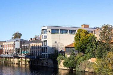 A riverfront view of buildings, including a multi-story beige structure with large windows, and older brick buildings, lined with trees under a clear blue sky in York, UK.
