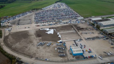 Aerial view of a large industrial site featuring rows of parked cars, construction equipment, and several warehouse buildings under a cloudy sky  at Copart yard, York, UK.