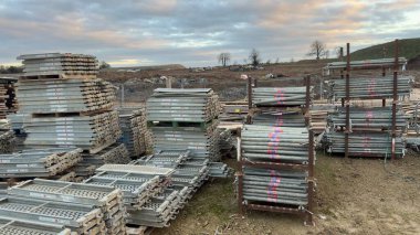 İnşaat alanında yığılı metal iskeleler ve borular var. Gökyüzü bulutlu ve gri.