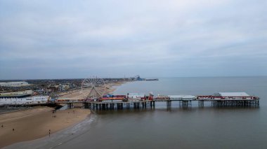 Havadan bakıldığında, Blackpool 'da bulutlu bir gökyüzü altında deniz kenarı iskelesi, dönme dolap, lunapark araçları ve sakin su üzerinde uzanan binalar görülüyor..