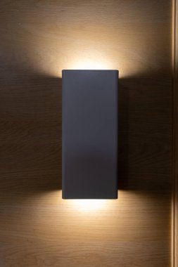 Modern, koyu gri dikdörtgen bir duvar lambası ahşap bir duvara monte edilir, yukarı ve aşağı doğru sıcak bir ışık yayar..