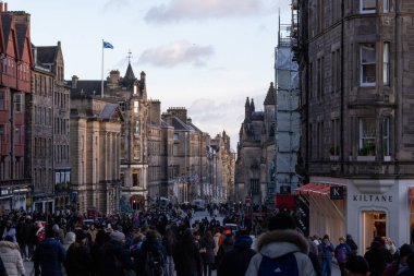 İnsanların alışveriş yaptığı ve bulutlu bir gökyüzünün altında yürüdüğü tarihi taş binaların olduğu kalabalık bir sokak manzarası. İskoçya 'nın Edinburgh şehrinde bir İskoç bayrağı dalgalanıyor.