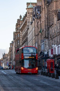 Kırmızı çift katlı bir otobüs tarihi taş binalar, kırmızı bir telefon kulübesi ve İskoçya 'nın Edinburgh şehrinde yaya trafiği olan kaldırım taşlı bir caddeden geçiyor..