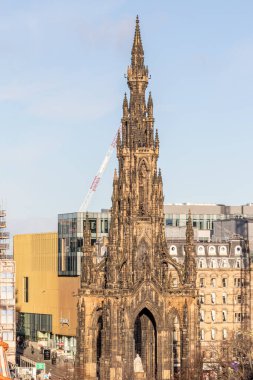 Ön planda karmaşık ayrıntılara sahip uzun, Gotik tarzı bir anıt yer alıyor. Arkasında, Edinburgh, İskoçya 'da soluk mavi bir gökyüzünün altında, modern sarı bir bina ve kemerli camlı daha eski bronz binalar görülebilir..