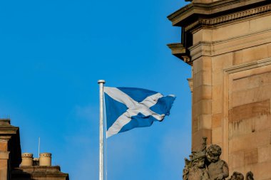 Bir İskoç bayrağı bayrak direğinde parlak mavi bir gökyüzüne doğru dalgalanıyor. Süslü kumtaşı mimarisi ve sağ tarafından görülebilen melek heykelleri var..