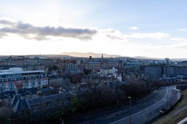 İskoçya 'nın Edinburgh kentindeki uzak tepelerde ve ön planda dolambaçlı bir yol bulunan bulutlu bir gökyüzünün altında genişleyen bir şehir..