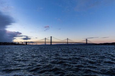 Geniş bir asma köprü koyu mavi su boyunca uzanır alacakaranlık gökyüzünün altında solda kara bulutlar ve sağda açık renkli bulutlar..