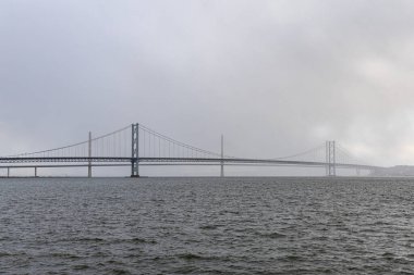 Uzun bir asma köprü, puslu, bulutlu bir gökyüzü altında geniş bir kara su genişliği boyunca uzanır, ikinci, daha küçük bir köprü uzaklıktan görülebilir..