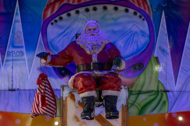 Parlak ışıklandırılmış, yakın plan çekimde canlı, karlı dağları olan çizgi film arkaplanı karşısında oturan bir Noel Baba figürü görülüyor..