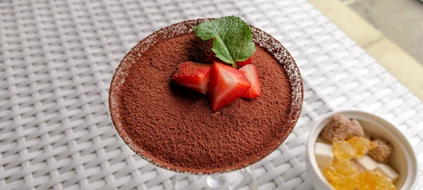 İştah açıcı bir tatlı. Tiramessu, çikolata serpiştirilmiş ve çilek dilimleri ve nane yaprağı ile süslenmiş, masanın üzerinde cam bir kasede. Metin için yer 