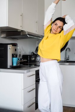 Brunette girl with bluetooth headphones dances in the kitchen in the morning and makes coffee in a coffee machine