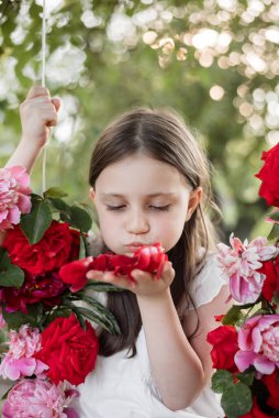 Çiçekler, güller ve şakayıklarla süslenmiş, salıncakta sallanan 5 yaşında küçük bir kızın portresi. Çiçek yapraklarına üfleyen kız. Bahar ve yaz konsepti