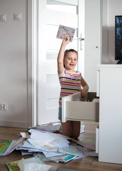 A preschool girl tidies up the dresser in her room. A child among a mess of a heap of papers, sheets and drawings