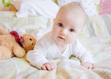 6 aylık bir bebek, karnının üstünde, oyuncak bir ayının yanında yatıyor. Yatağında oyuncağı olan bir kız.