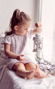 Pencere pervazında elinde çelenk ve el feneri olan küçük bir kız oturuyor. Pastel, renkli görüntü. Noel ve Yeni Yıl konsepti. Karantina ve pandemik konsept.