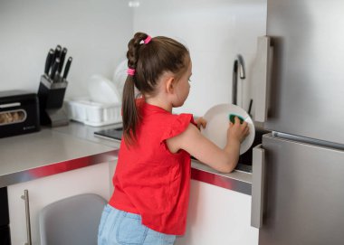 Anaokulu kızı evde bulaşıkları yıkıyor. Çocuk bulaşık yıkamada ebeveynlere yardım eder, çocuklar için ev işi yapar..