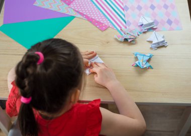 Anaokulundan bir kız, origami kurbağası ve kağıttan bir tekne yapar, çocuklar için yaratıcılık, karantinadaki çocuklar için etkinlikler. İzolasyon, koronavirüs 2021.