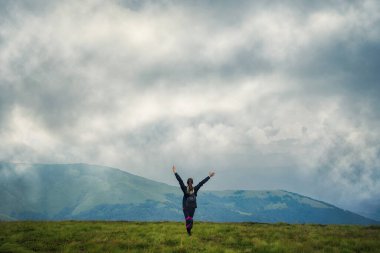 Sırt çantalı ve dağlarda elini kaldıran bir kız. Alçak bulutlar. Sis. Hedefe ulaşma başarısı. Başarı. Turizm. Seyahat.
