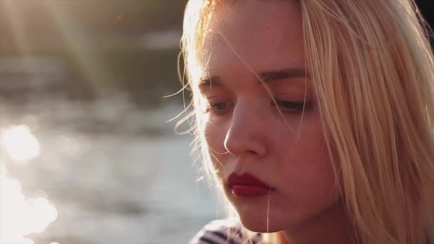 Menina caucasiana nova bonita com cabelo loiro e lábios vermelhos no pôr do sol na frente da água. A olhar para a câmara. Retrato, tiro de perto. Movimento lento — Vídeo de Stock