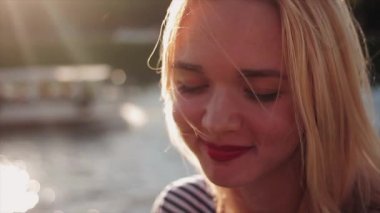 Güzel genç beyaz kızla sarı saçları ve günbatımı üzerinde su önünde oturan kırmızı dudaklar gülümsüyor. Kameraya bakıyor. Portre, yakından atış. Slowmotion. Tarafından geçen tekne