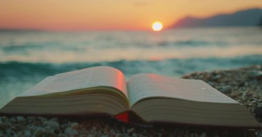 Açık kitap kumsalda çakıl taşları üzerinde uzanır ve alacakaranlıkta deniz dalgaları vardır..