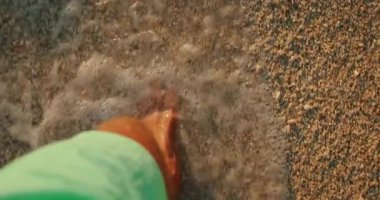 Şortlu adam gün batımında beyaz çakıl taşlı sahilde denizin köpüklü suyuna adım atar..