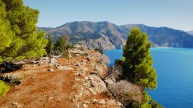 Yunanistan 'da deniz ve dağ manzaralı kayalık kıyı şeridi boyunca yürüyen yürüyüşçü kadın.