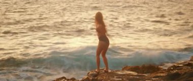 Siyah mayo giymiş bir kadın altın saat ışığında denizin kenarında dans ediyor..