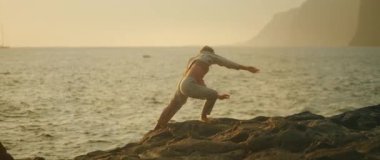 Dansçı, doğaçlama bir anda gün batımında kayalık sahilde eğilir..